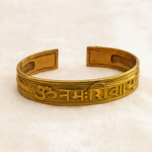 May include: A gold-coloured cuff bracelet with engraved Sanskrit text. The open-ended bracelet has a rectangular design on the ends and a detailed pattern around the band. The text is centered on the bracelet.