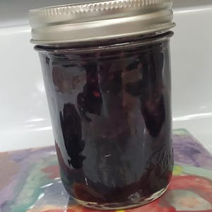 May include: A glass jar filled with dark purple jam, sealed with a silver lid. The jar sits on a colorful, abstract patterned surface. The jam appears thick and glossy, suggesting a homemade preserve.