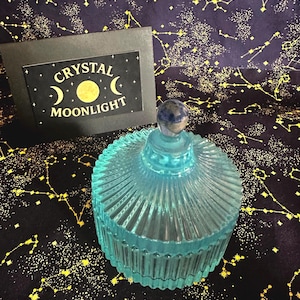 May include: A turquoise glass jar with a ribbed design and a matching lid. The lid is topped with a small, round, blue stone. A black card with the words "CRYSTAL MOONLIGHT" is in the background.