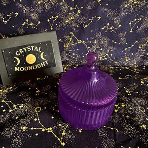 Puede incluir: Un tarro de vidrio morado acanalado con tapa y asa esférica. El tarro está sobre una tela azul oscuro estampada con constelaciones doradas. También hay un letrero negro con las palabras "Crystal Moonlight".