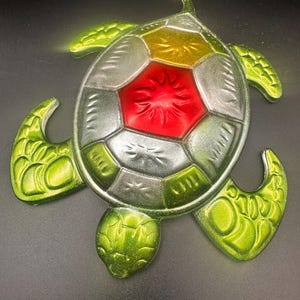 May include: A decorative turtle figurine with a segmented shell design. The shell features a red center, surrounded by silver, gold, and green sections. The turtle's head and flippers are a vibrant green, creating a colorful, eye-catching piece.