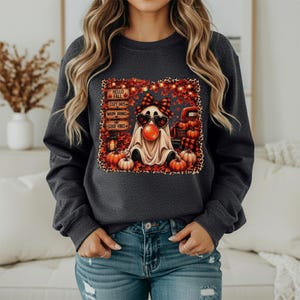 May include: A dark gray crewneck sweatshirt featuring a graphic print of a ghost wearing heart-shaped sunglasses and a plaid bow. The design includes pumpkins, autumn leaves, a red truck, and text that reads HELLO FALL, COZY DAYS, WARM DRINKS, AND GOOD VIBES.