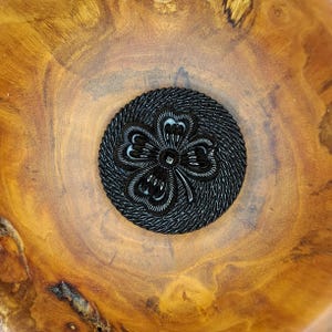 Puede incluir: Un botón negro circular que presenta un diseño de trébol de cuatro hojas en relieve en el centro. El botón tiene un borde texturizado de estilo trenzado que rodea el motivo del trébol, sobre una superficie de madera pulida y lisa.