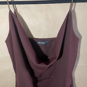 May include: A burgundy-colored dress with spaghetti straps and a cowl neckline. The dress is hanging on a black hanger. The tag reads "PRETTYLITTLETHING".