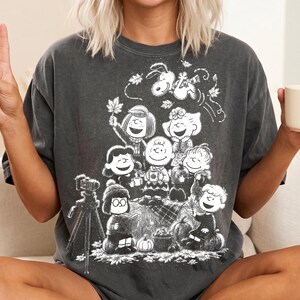 May include: A charcoal grey short-sleeve t-shirt featuring a white graphic print of the Peanuts characters. The design depicts Charlie Brown, Snoopy, and friends gathered outdoors with pumpkins, autumn leaves, and a camera on a tripod stand.