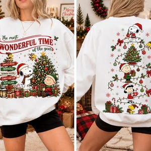 May include: A white crewneck sweatshirt featuring Peanuts characters. The front displays Snoopy and Charlie Brown near a Christmas tree with signs reading Santa's House, North Pole, Reindeer Crossing, Candy Cane Lane, and Elf Village. The back shows a tree design with Snoopy, Charlie Brown, Lucy, and Woodstock.