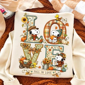 May include: A cream-colored sweatshirt featuring the word LOVE in large letters decorated with autumn themes. Snoopy and Woodstock are illustrated throughout, wearing seasonal hats and scarves, surrounded by pumpkins, sunflowers, and leaves. The text FALL IN LOVE is printed on a banner at the bottom.