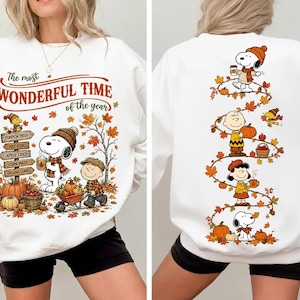 May include: A white long-sleeve sweatshirt featuring Peanuts characters in autumn scenes. The front displays Snoopy, Charlie Brown, and text reading The most WONDERFUL TIME of the year. The back shows a vertical design of characters with falling leaves.