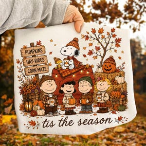 May include: A white sweatshirt featuring a Peanuts graphic with Snoopy, Woodstock, and friends in an autumn setting. The design includes a wooden sign reading PUMPKINS, HAY-RIDES, CORN MAZE, and the text tis the season, surrounded by pumpkins and fall foliage.