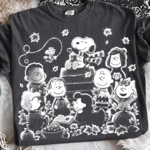 Può includere: Una t-shirt grigio antracite con una stampa grafica bianca dei personaggi dei Peanuts che celebrano l'autunno. Il design include Snoopy, Charlie Brown e amici circondati da foglie cadenti, zucche, un libro e un cesto di mele.