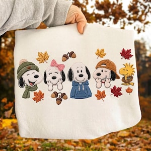 May include: A white sweatshirt featuring an embroidered design of four cartoon dogs and a small yellow bird. The characters are wearing autumn accessories like a green beanie, a pink bow, a blue hoodie, and a brown cap, surrounded by maple leaves and acorns.
