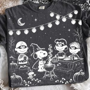 以下が含まれることがあります： ハロウィンの仮装をしたピーナッツのキャラクターたちが描かれた、チャコールグレーのTシャツ。白い線画で、スヌーピー、チャーリー・ブラウン、仲間たちがキャンプファイヤーを囲む様子、ストリングライト、三日月、カボチャがデザインされています。