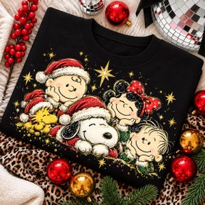 May include: A black crewneck sweatshirt featuring a graphic print of Charlie Brown, Snoopy, Woodstock, Lucy, and Linus wearing Santa hats. The characters are surrounded by gold stars and sparkles on a dark background.