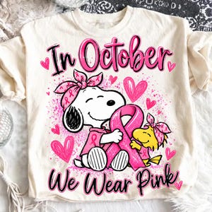 Puede incluir: Una camiseta de color crema con un gráfico de Snoopy y Woodstock con diademas y camisas rosas, abrazando un gran lazo rosa de concienciación sobre el cáncer de mama. El texto In October We Wear Pink está rodeado de corazones y salpicaduras.