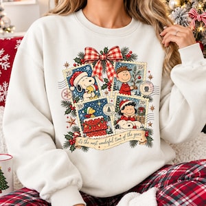 Peut inclure: Un sweat-shirt blanc à col rond orné d'un motif représentant quatre timbres illustrés avec les personnages Peanuts dans des scènes de Noël, surmonté d'un nœud vichy rouge et blanc et du texte, "it's the most wonderful time of the year."