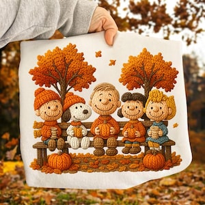 Puede incluir: Una sudadera blanca con un gráfico de estilo tejido y texturizado de los personajes de Peanuts sentados en un banco de madera bajo dos árboles de otoño. Los personajes sostienen calabazas, rodeados de hojas caídas y follaje naranja.