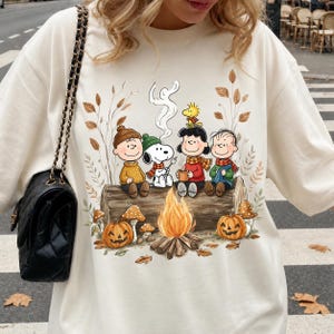 以下が含まれることがあります： クリーム色のTシャツ。チャーリー・ブラウン、スヌーピー、ウッドストック、ルーシー、ライナスが焚き火のそばの丸太に座っているグラフィックプリントが施されています。秋の葉、キノコ、2つのジャック・オー・ランタンが描かれた秋をテーマにしたデザインです。