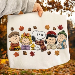 May include: A white sweatshirt featuring an embroidered design of the Peanuts characters including Snoopy, Woodstock, Charlie Brown, Sally, Lucy, and Linus wearing autumn scarves and hats, surrounded by colorful embroidered fall leaves and acorns.