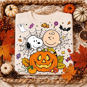 May include: A cream-colored sweatshirt featuring a graphic print of Snoopy and Charlie Brown behind a large orange jack-o'-lantern. The design includes a spider web, a white ghost, a black bat, colorful candies, and autumn leaves.