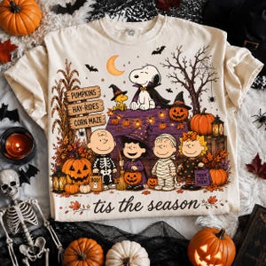 May include: A cream-coloured t-shirt featuring a Peanuts Halloween graphic with Snoopy, Charlie Brown, and friends in costumes. The design includes a signpost reading PUMPKINS, HAY-RIDES, CORN MAZE, a crescent moon, and the text tis the season.