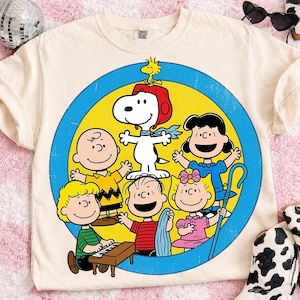以下が含まれることがあります： ピーナッツのキャラクターが描かれた円形のグラフィックが特徴のクリーム色のTシャツ。スヌーピー、チャーリー・ブラウン、ルーシー、ライナス、サリー、シュローダーが青い縁取りの黄色い円の中に配置されており、かすれたプリントスタイルが施されています。