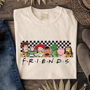 Può includere: Una t-shirt color crema a girocollo con una grafica dei personaggi di Toy Story Woody, Buzz Lightyear, Jessie, Mr. Potato Head, Rex, Hamm e Slinky Dog davanti a un motivo a scacchi bianco e nero con la parola FRIENDS stampata sotto.