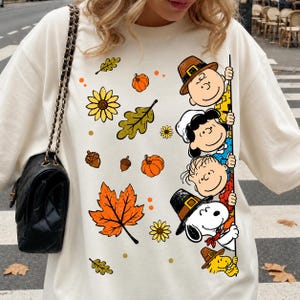 May include: A cream-colored crewneck sweatshirt featuring a graphic print of Peanuts characters including Charlie Brown, Lucy, Linus, Snoopy, and Woodstock. The design includes autumn-themed elements like orange pumpkins, acorns, and fall leaves.