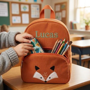 May include: Orange corduroy backpack with a fox face design on the front pocket. The backpack has the name "Lucas" embroidered in green. The front pocket is open, revealing colored pencils and a brush. A hand is putting a pencil case into the pocket.