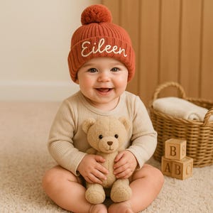 Op de afbeelding: Een lachende baby draagt een roestkleurige gebreide muts met de naam "Eileen" in het wit geborduurd. De baby houdt een beige teddybeer vast en draagt een beige onesie. Houten alfabetblokken en een rieten mand staan op de achtergrond.