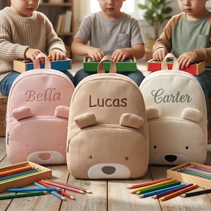 May include: Three corduroy backpacks in pink, tan, and cream, each with a bear face design and embroidered name. The pink backpack says "Bella", the tan backpack says "Lucas", and the cream backpack says "Carter". Colored pencils and wooden toy blocks are also visible.