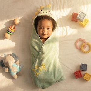 May include: A baby wrapped in a light green hooded towel with a dinosaur design. The towel has the name "Lily" embroidered in yellow. Toys, including a wooden rattle, a plush elephant, and colorful blocks, surround the baby.