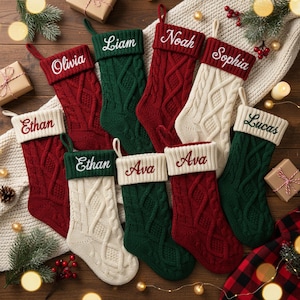 May include: A collection of personalized Christmas stockings in various colors, including red, green, and cream. Each stocking features a cable knit design and a name embroidered in white or red thread. The names include Olivia, Liam, Noah, Sophia, Ethan, Ava, and Lucas.