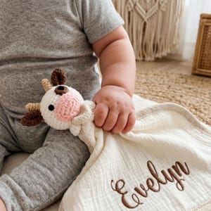 May include: A white and brown crochet cow toy with a pink nose and brown horns is held by a baby. The baby is also holding a cream-colored muslin blanket with the name "Evelyn" embroidered in brown.