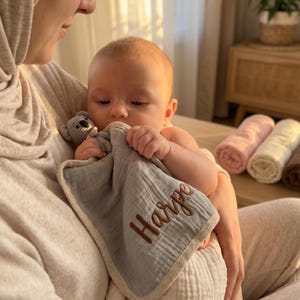 May include: A baby is held, clutching a gray muslin security blanket with the name "Harpe" embroidered in brown. A small gray knitted teddy bear is also held. Rolled-up blankets in pink, cream, and brown are in the background.