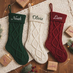 Puede incluir: Tres calcetines navideños personalizados en verde, crema y rojo, cada uno con un diseño de punto trenzado. Los nombres "Noah", "Olivia" y "Liam" están bordados en los puños. Los calcetines se exhiben sobre una manta blanca con regalos envueltos.