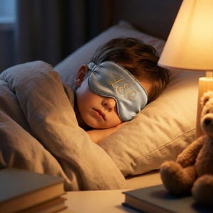 May include: A sleeping mask with the name "LEO" embroidered in gold on a light blue satin fabric. The mask is worn by a person lying in bed, with a pillow and blanket. A lamp and teddy bear are in the background.