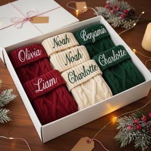 Puede incluir: Una caja de regalo blanca contiene seis calcetines navideños personalizados en rojo, crema y verde. Cada calcetín está bordado con un nombre en un color complementario. La caja está decorada con vegetación festiva y una vela encendida.