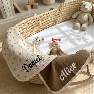Könnte beinhalten: Zwei Baby-Schmusetücher mit gestickten Namen, eines beige mit braunen Herzen und dem Namen "Daniel", das andere braun mit "Alice". Ein geflochtener Korb enthält die Decken, mit einem gestrickten Bären und einem Fuchspielzeug.
