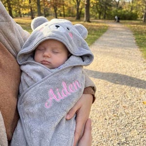 May include: A sleeping baby wrapped in a grey hooded towel with a bear face design. The towel is embroidered with the name "Aiden" in pink script. The baby is being held outdoors on a sunny day.
