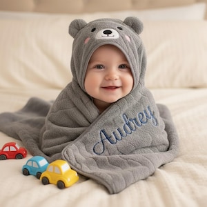 May include: A smiling baby wrapped in a gray hooded towel with bear ears. The towel is embroidered with the name "Audrey" in blue script. Three toy cars in red, blue, and yellow are in the foreground.