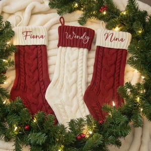 May include: Three cable-knit Christmas stockings in red and cream, each with a white cuff embroidered with a name: Fiona, Wendy, and Nina. The stockings are surrounded by evergreen branches and lights, creating a festive holiday scene.