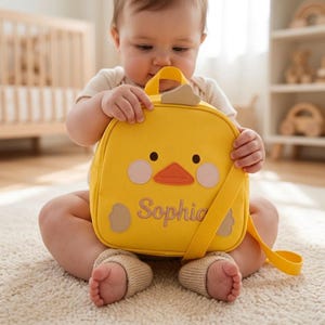 May include: A yellow backpack with a chick design, featuring black eyes, an orange beak, and light pink cheeks. The name "Sophie" is embroidered on the front. The bag has a top handle and a shoulder strap.