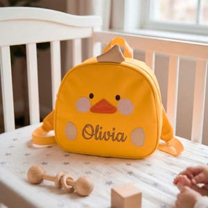 May include: A yellow children's backpack shaped like a duck. It has a beak, eyes, and cheek details. The name "Olivia" is embroidered on the front. The bag has adjustable straps and a top handle.