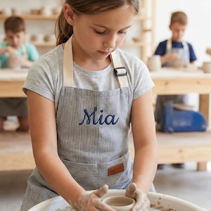 Może przedstawiać: Młoda osoba w szarej, pasiastym fartuchu z wyhaftowanym na niebiesko imieniem "Mia". Fartuch ma małą brązową skórzaną metkę z napisem "apron". Osoba pracuje z gliną na kole garncarskim. Inne osoby są w tle.