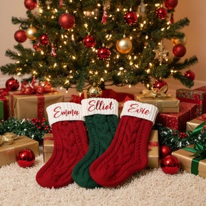 May include: Three personalized Christmas stockings in red and green, with names embroidered in cursive. The stockings are cable-knit and sit in front of a decorated Christmas tree with ornaments and wrapped gifts.