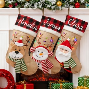 Könnte beinhalten: Drei personalisierte Weihnachtsstrümpfe mit den Namen Amelia, James und Clara. Jeder Strumpf hat ein Jute-Design mit einer rot-schwarz karierten Manschette. Die Strümpfe sind mit festlichen Figuren dekoriert: einem Bären, einem Schneemann und dem Weihnachtsmann.