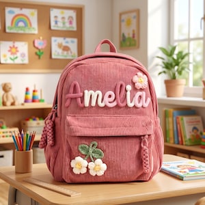 Peut inclure: Un sac à dos en velours côtelé rose avec le nom "Amelia" en tricot. Le sac à dos comporte une poche avant, une poignée supérieure et des appliqués de fleurs et de cerises. Il est posé sur un bureau en bois dans une salle de classe.