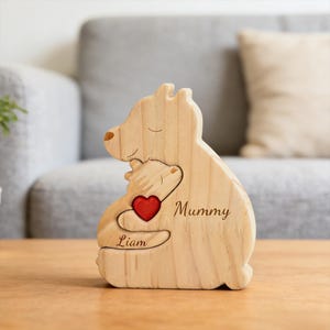 May include: A light-colored wooden bear figurine featuring a larger bear embracing a smaller bear. The larger bear is engraved with the word Mummy, and the smaller bear is engraved with the name Liam. A small, bright red wooden heart is attached.