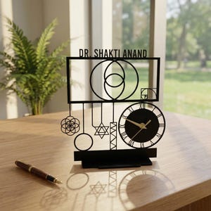 May include: Black laser-cut desk clock with the text "DR. SHAKTI ANAND". The clock features a circular face with Roman numerals and gold hands. Decorative geometric shapes and symbols are incorporated into the design.