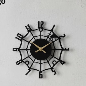 May include: Black spiderweb-shaped wall clock with gold hands and a black clock face. The clock features a spider design and is mounted on a white wall. The brand name "Lightinggo" is in the top left corner.